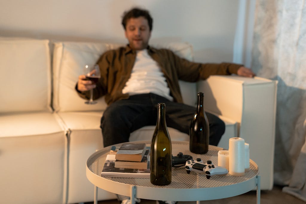 A man sits on a couch with wine bottles, a game controller, and books, expressing relaxation or contemplation.