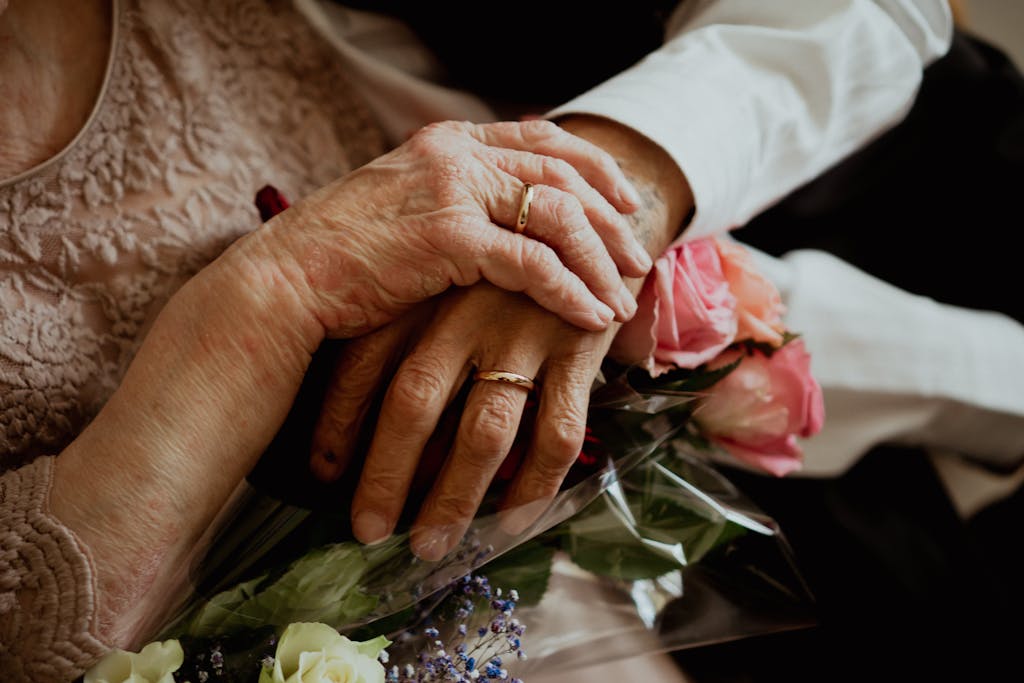 Close-up of elderly hands clasped together with wedding rings and a bouquet of roses, symbolizing love and commitment.